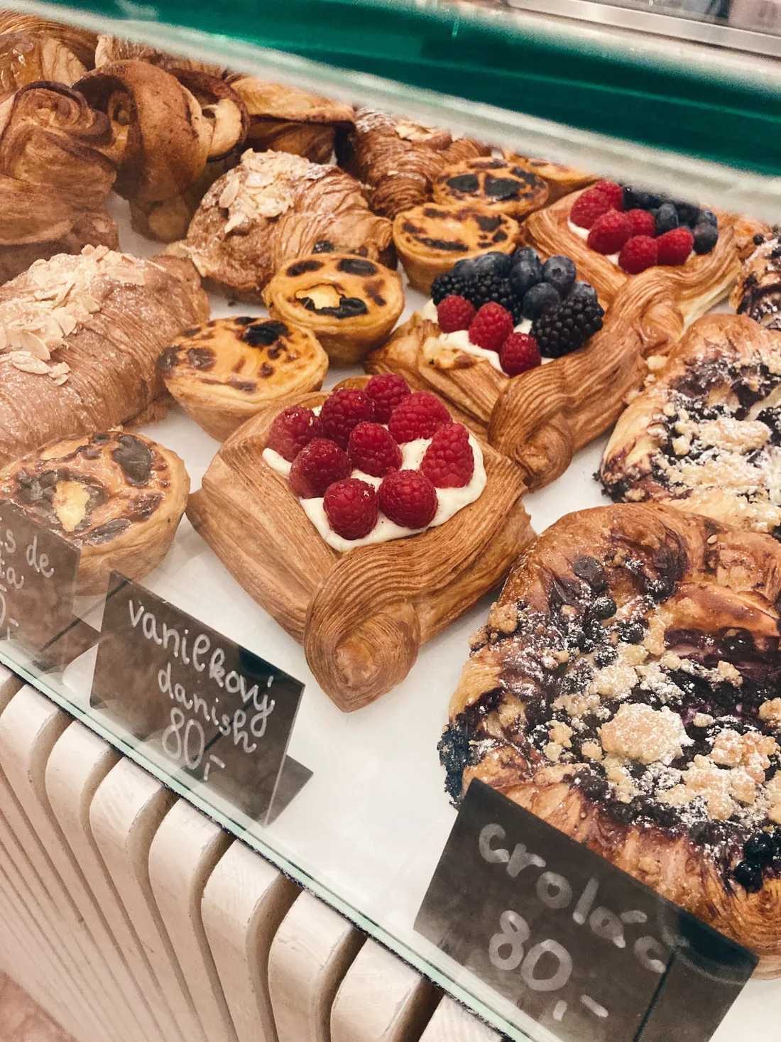 Vitrína s danishem, croissanty, croláčem a pastéis de nata s cenovkami v pekárně.