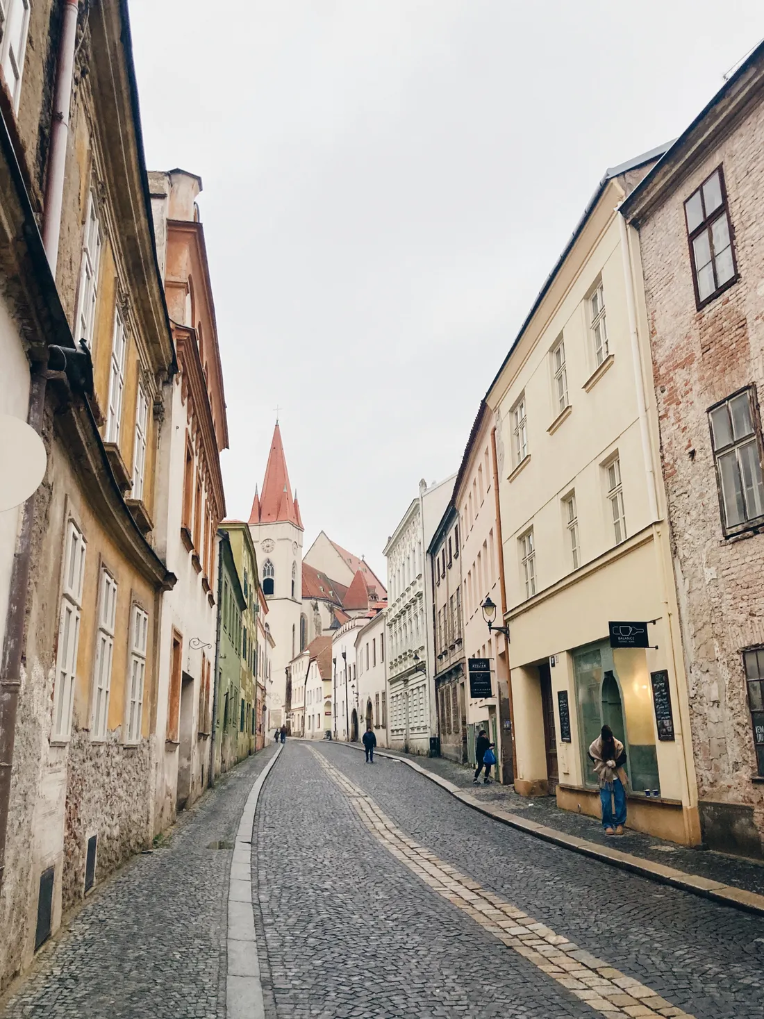 Úzká dlážděná ulice ve Znojmě s barevnými domy a kostelní věží s červenou střechou.