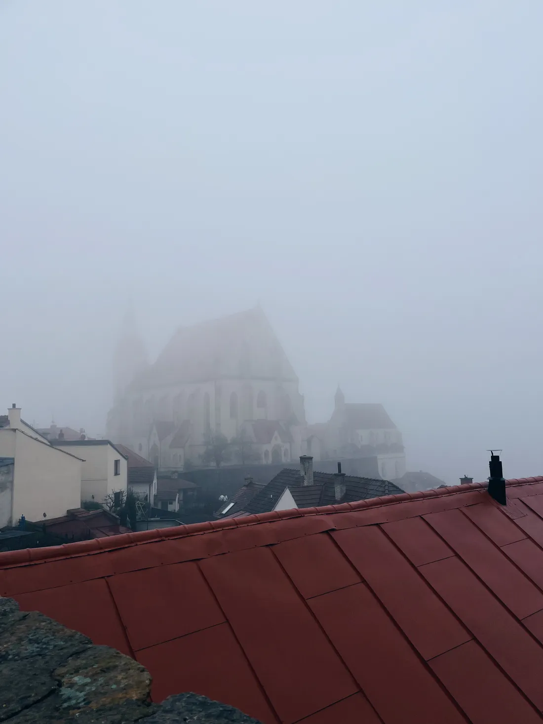 Pohled přes červenou střechu na město a gotický kostel zahalený v husté mlze.
