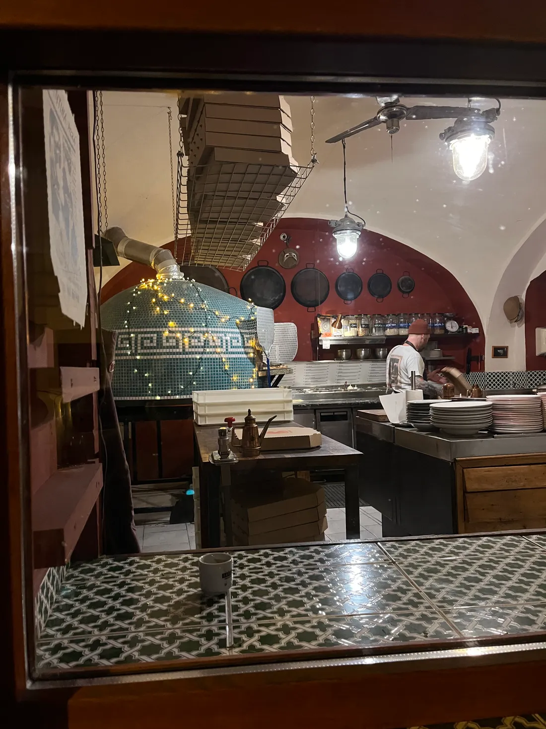 Interiér pizzerie s mozaikovou modrozelenou pecí, světýlky, pracovním pultem a hromadou talířů.