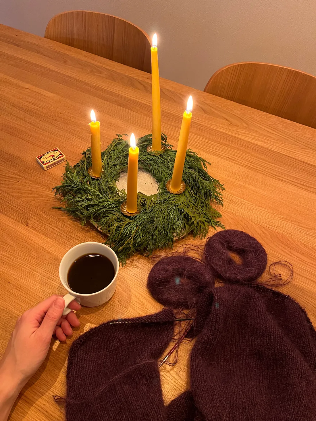 Zelený adventní věnec se čtyřmi zapálenými svíčkami, bílý hrnek kávy a růžovofialové pletení na dřevěném stole.