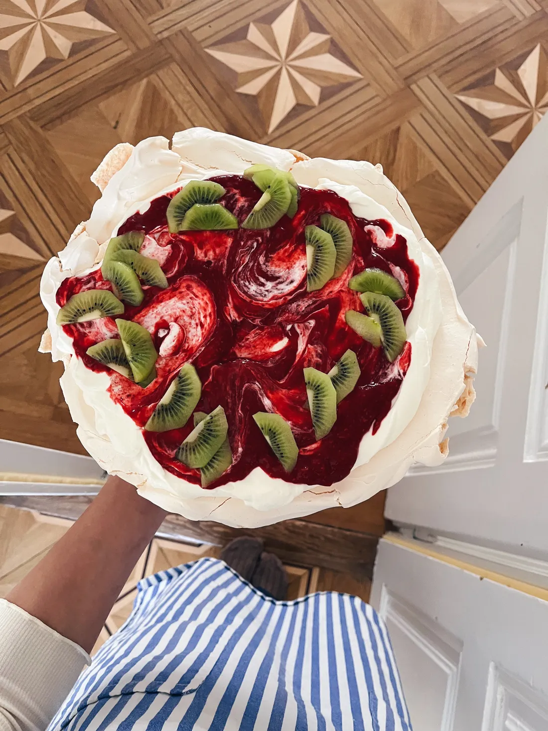 Pavlova se šlehačkou, červenou omáčkou a plátky kiwi držená v modrobílém pruhovaném zástěře nad parketami.