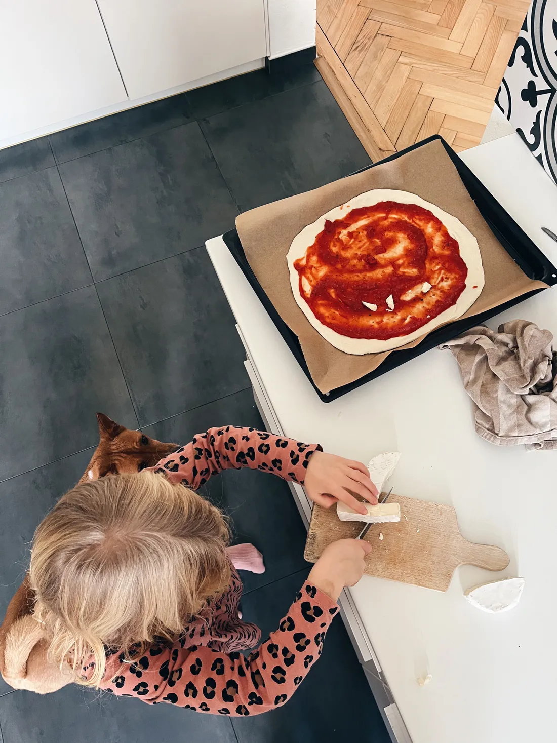 Dítě v leopardím tričku krájí sýr na prkénku vedle pizzy na plechu, pod stolem hnědý pes.