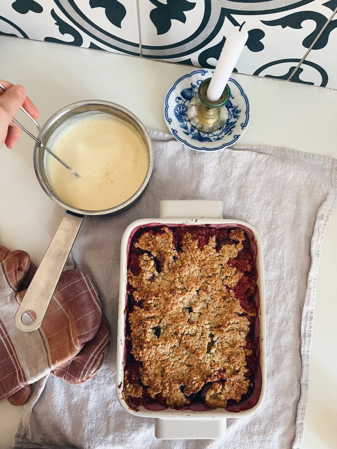 Ovocný crumble v bílé míse na šedé utěrce, vedle rendlík se světlou omáčkou a pruhovaná chňapka.