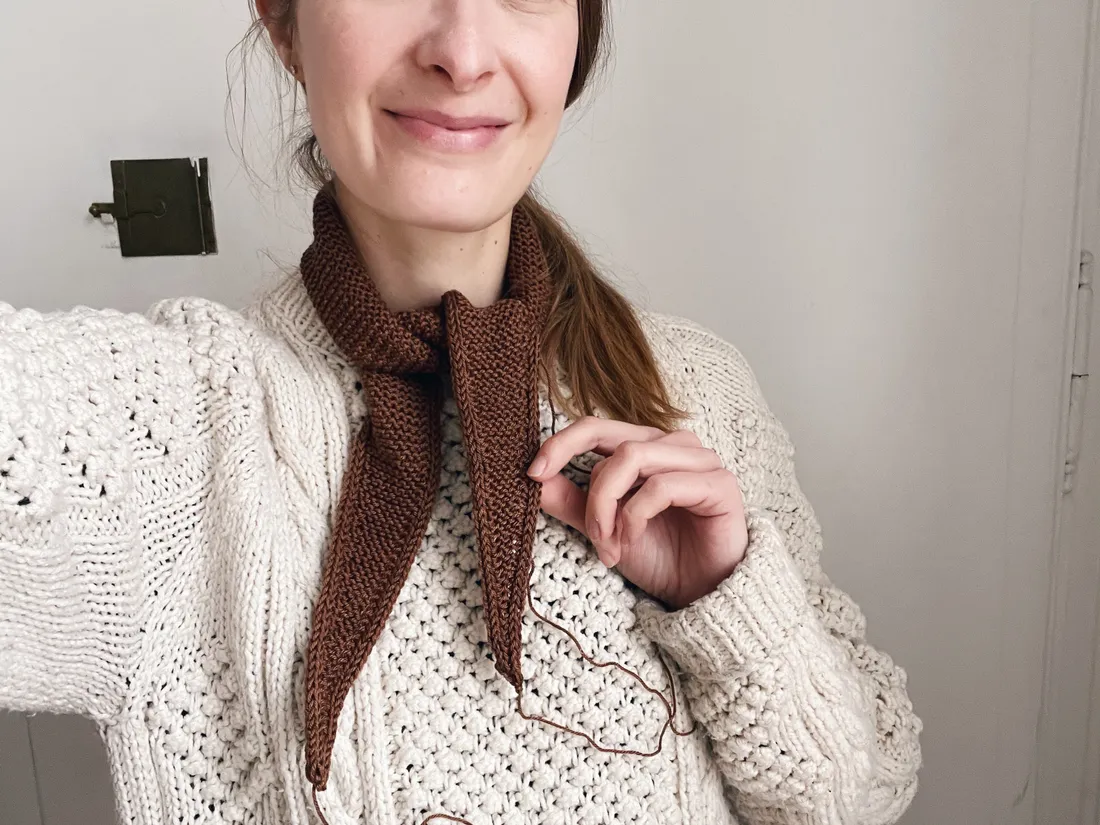 Žena v bílém svetru s ručně pleteným hnědým šátkem, drží konec příze.