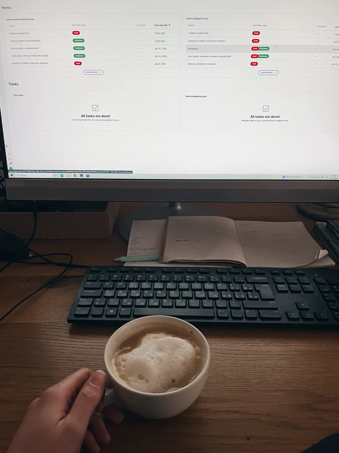 Ruka drží šálek kávy s mlékem, na monitoru přehled článků a úkolů Kentica.
