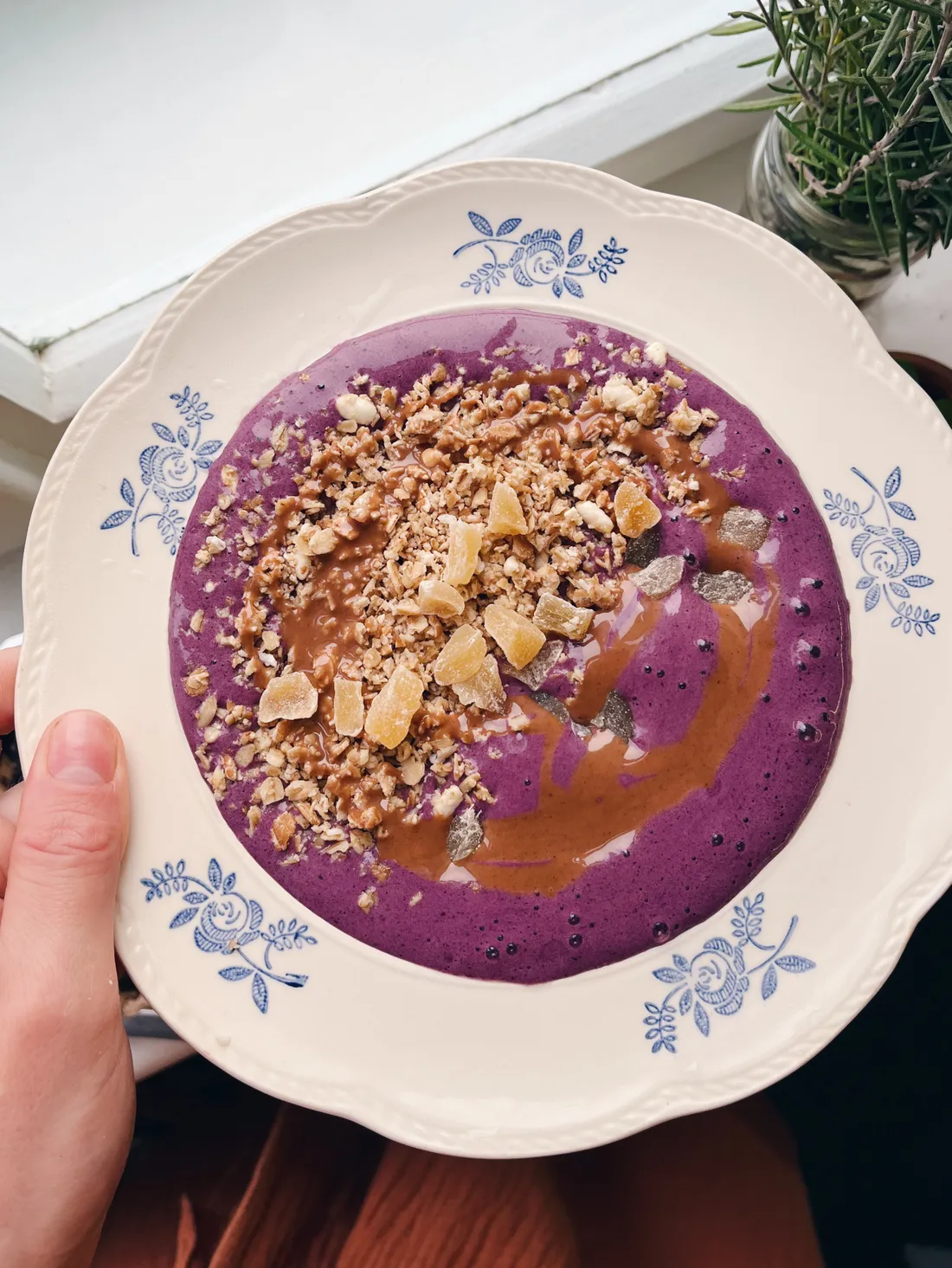 Fialová smoothie miska s granolou, ořechovým máslem a sušeným ovocem na zdobeném modrobílém talíři.