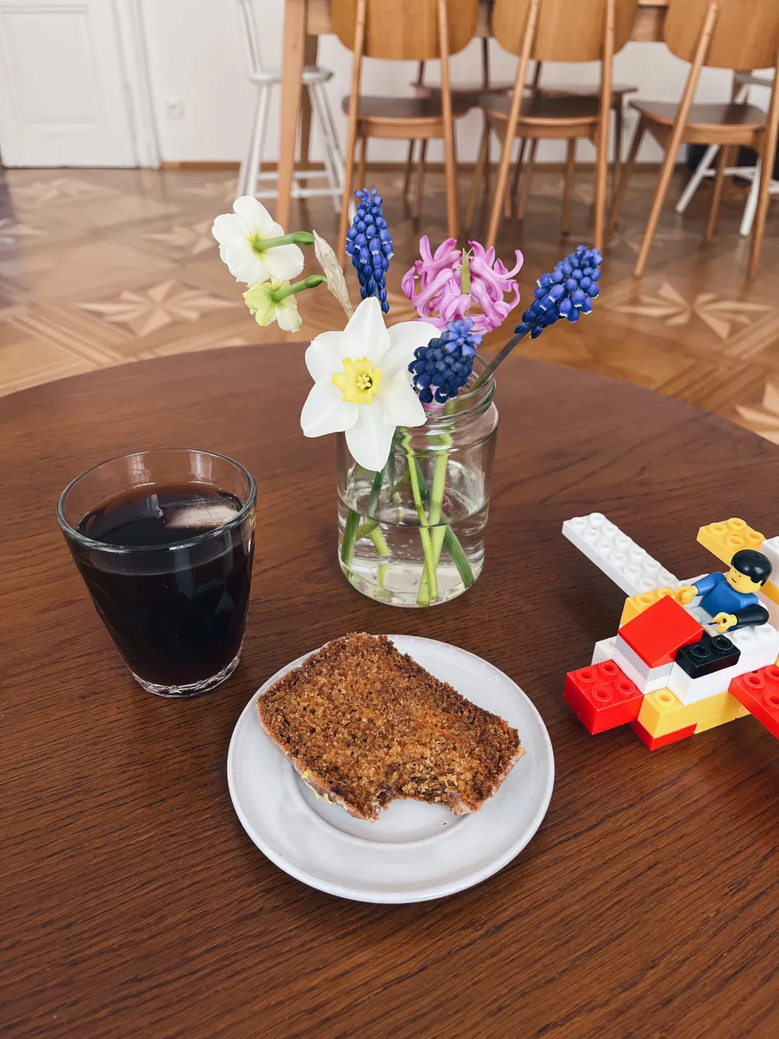 Sklenice koly, kousek chleba na talíři, kytice narcisů a hyacintů a lego letadlo na kulatém stole.