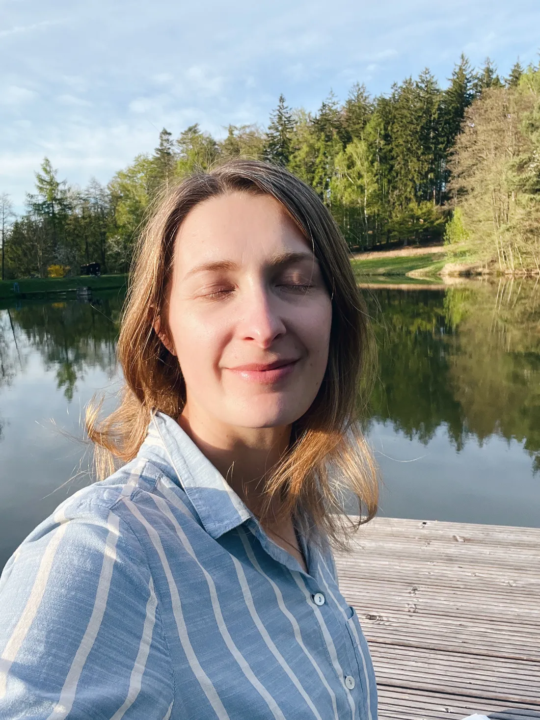 Žena se zavřenýma očima a úsměvem na dřevěném molu u klidného jezera se zrcadlícím lesem.
