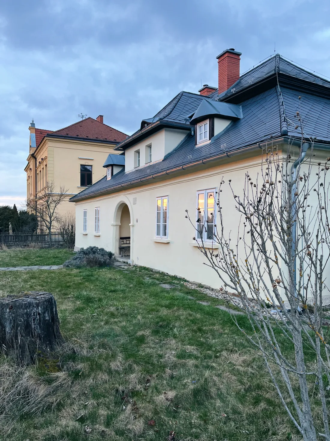 Krémový dům s šedou mansardovou střechou, v pozadí žlutá budova, trávník s pařezem pod mraky.