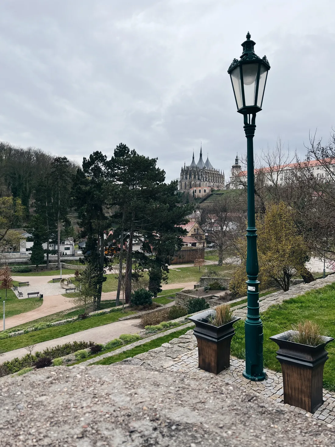 Pohled z terasy na park a chrám sv. Barbory, pouliční lampa a květináče pod oblohou.