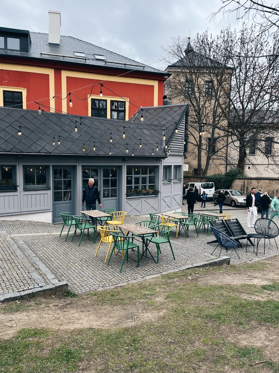 Venkovní posezení s barevnými židlemi a dřevěnými stoly na dlážděné terase, nad ním světelný řetěz.