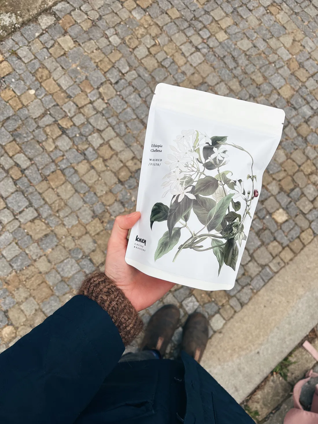 Ruka v tmavě modré bundě a hnědém pleteném svetru drží bílý sáček kávy Kmen s botanickou ilustrací květů na pozadí dlážděné ulice.