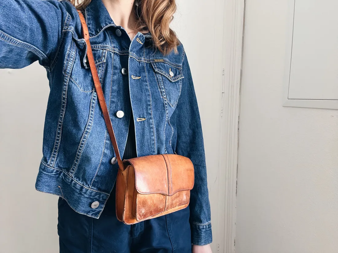 Žena v džínové bundě s hnědou koženou crossbody kabelkou na světlém pozadí.