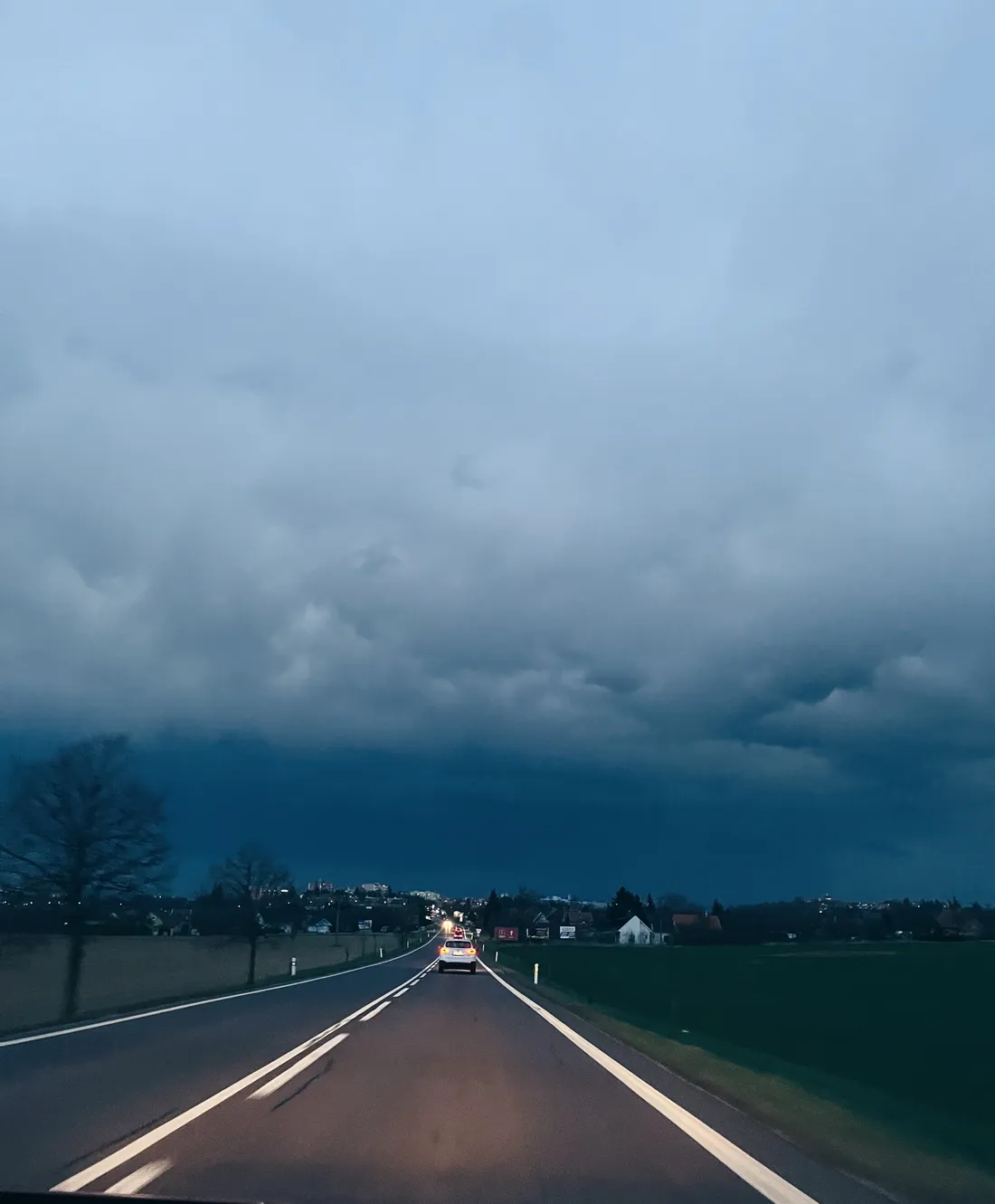 Silnice do města pod dramatickou bouřkovou oblohou, v dálce auto a světla.