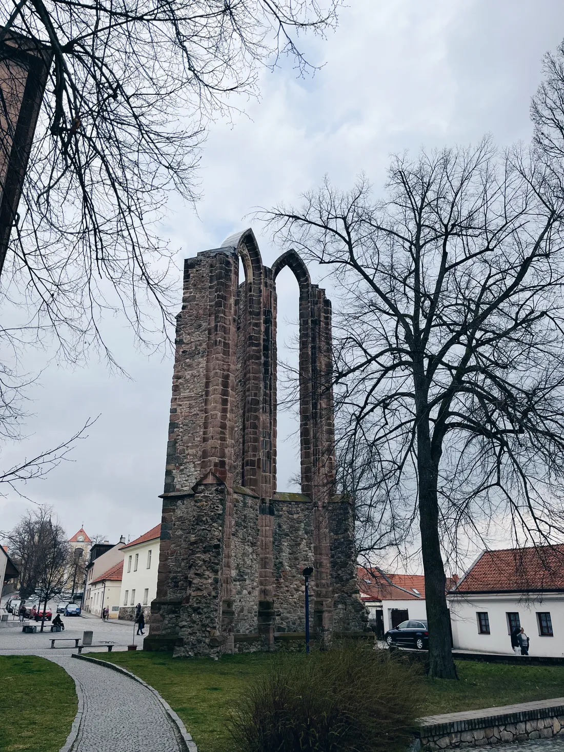 Vysoká kamenná gotická ruina se dvěma štíhlými lomenými oblouky v parku, v pozadí domy s červenými střechami.