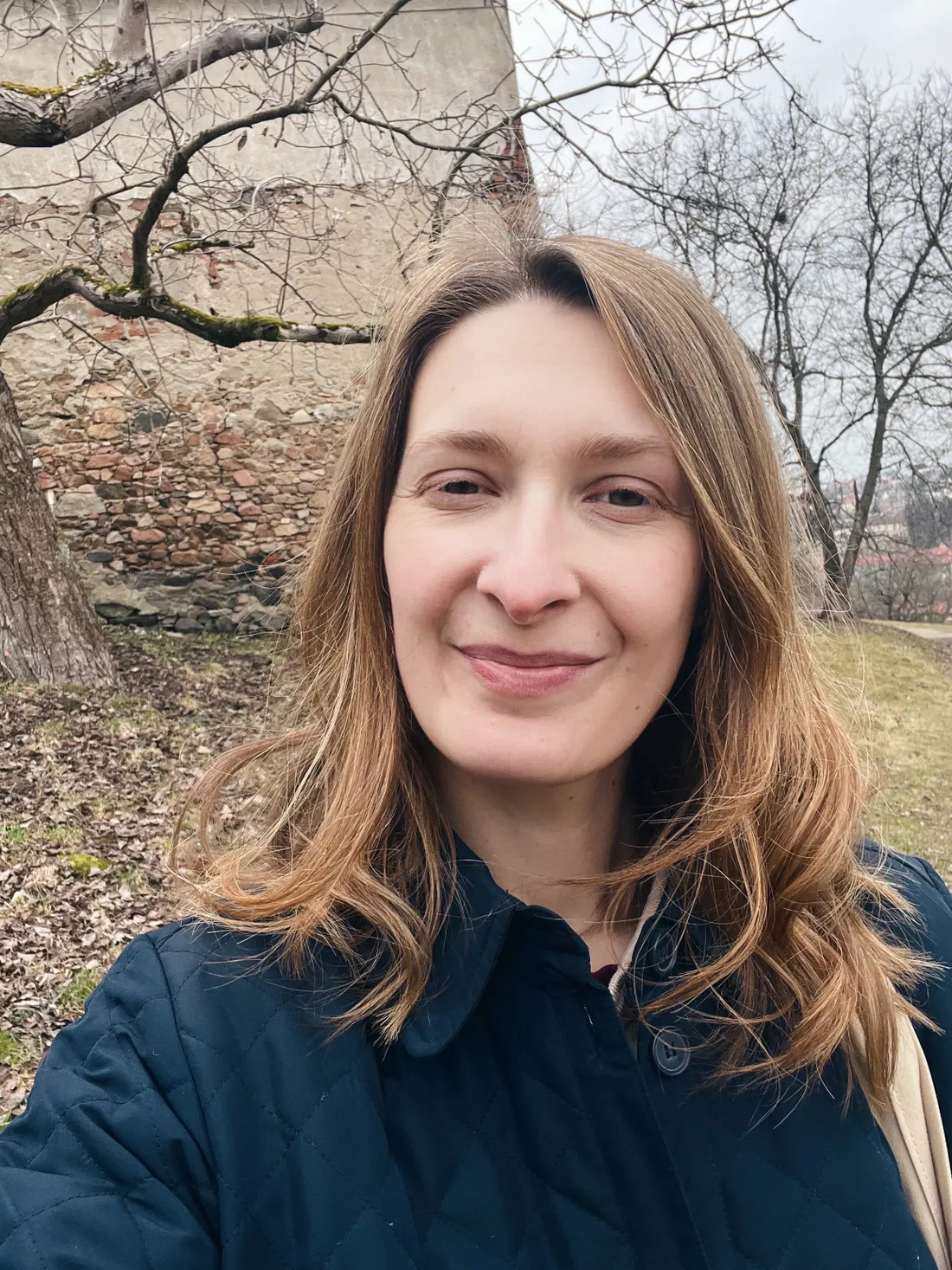 Usmívající se žena v tmavě modré prošívané bundě před kamennou zdí a holými stromy.