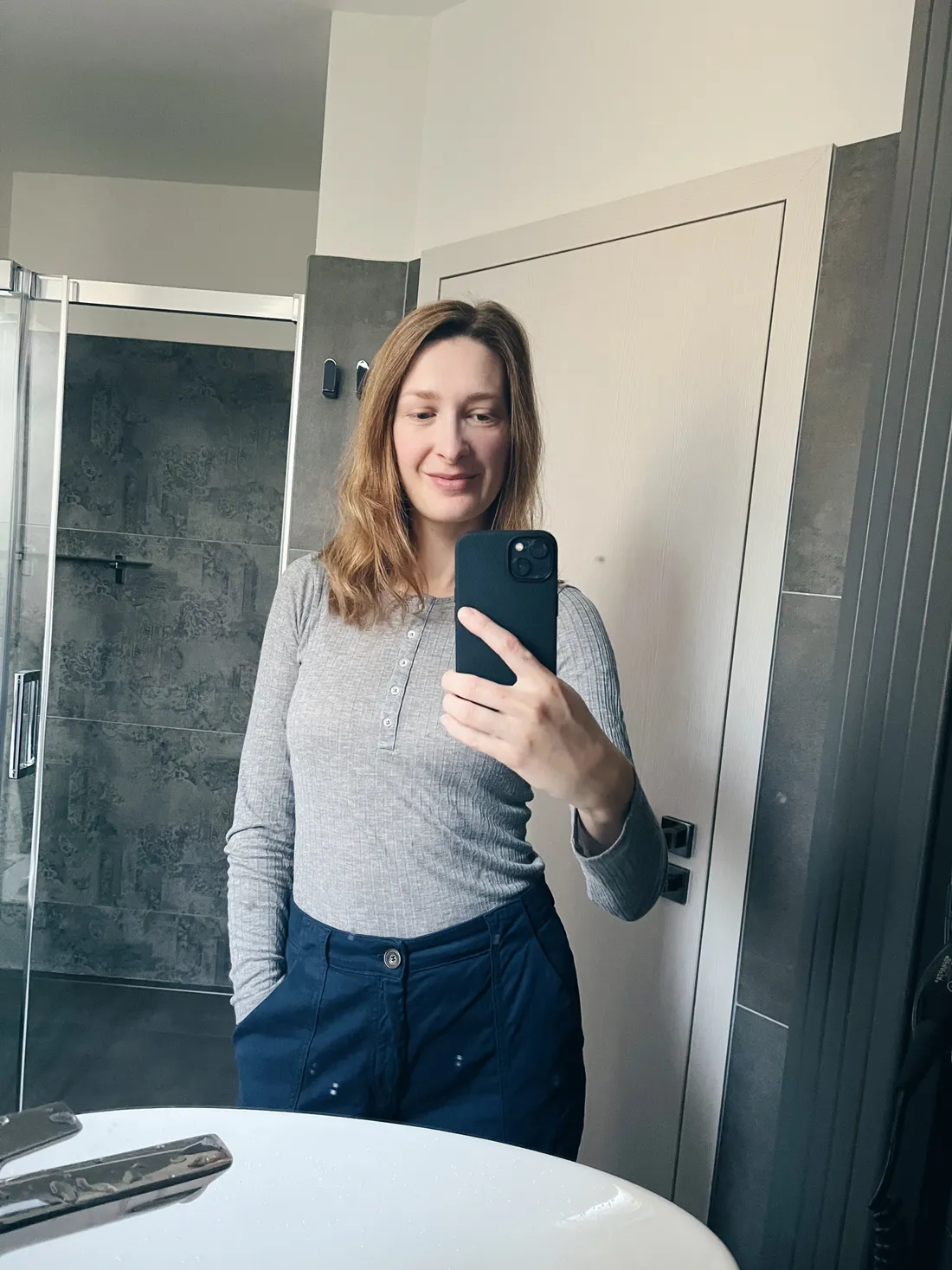 Selfie v koupelnovém zrcadle: žena v šedém pleteném svetru a tmavých kalhotách drží černý telefon u umyvadla.