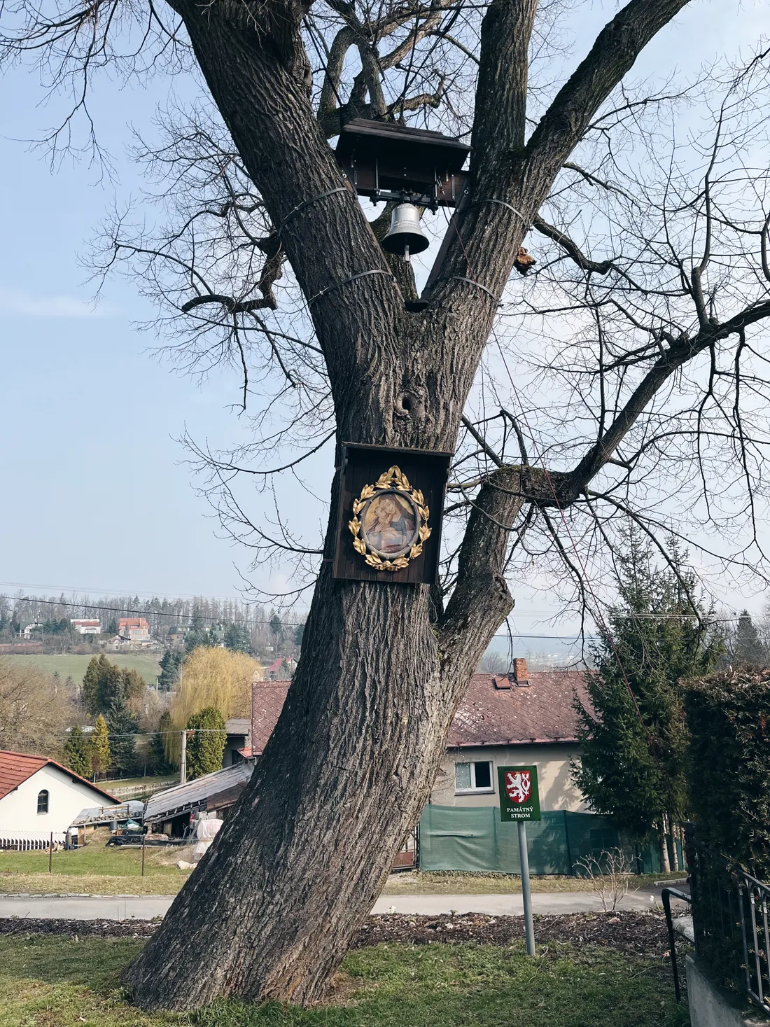 Památný strom se zvonicí a ikonou, zelená tabule v malé vesnici.