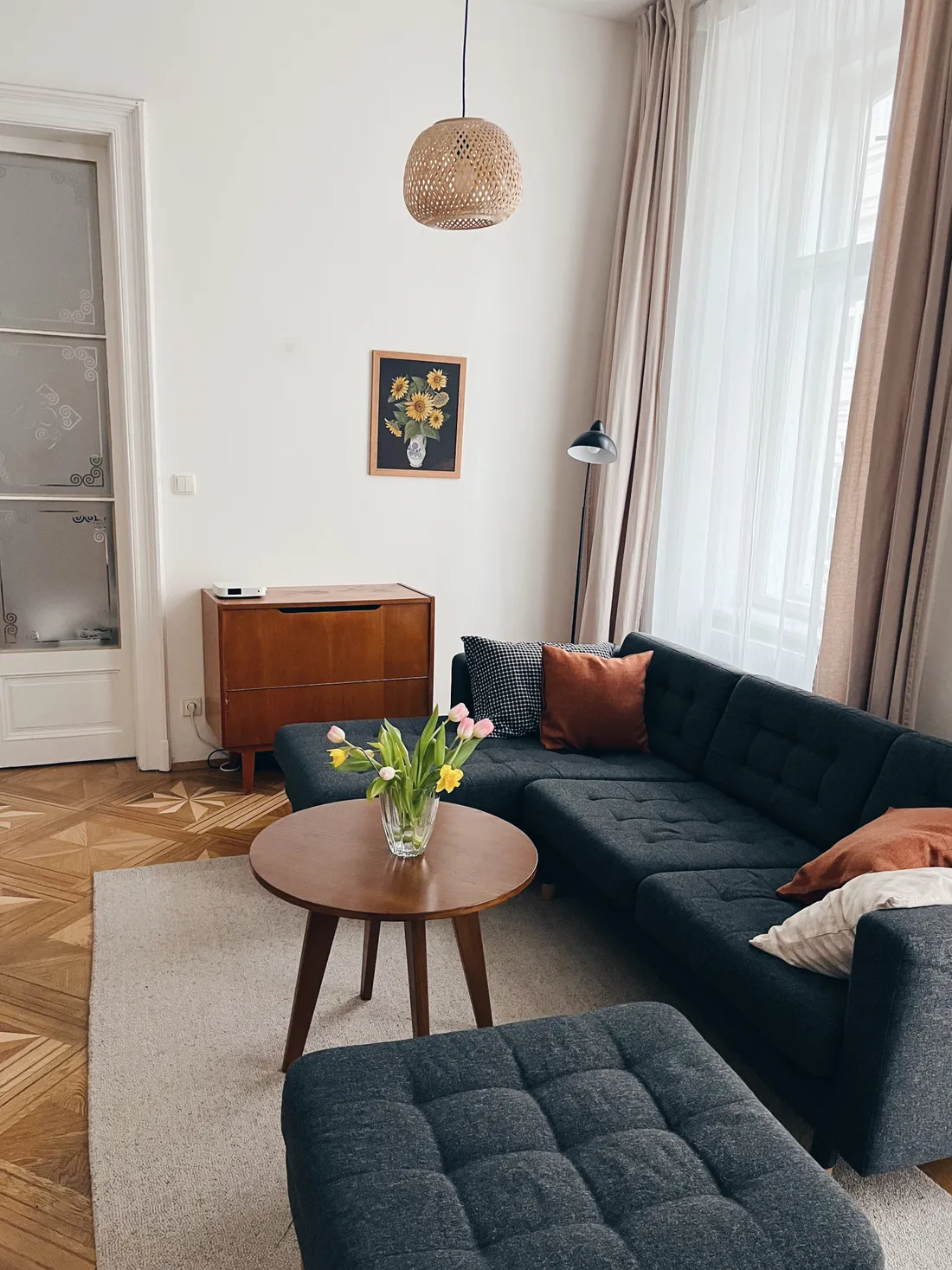 Obývací pokoj se šedou sedačkou, kyticí tulipánů na kulatém stolku a parketami s hvězdicovým vzorem.