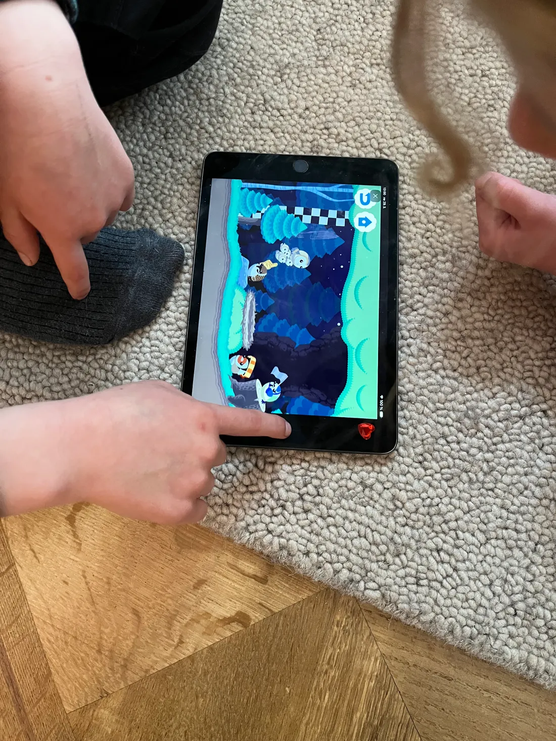Děti hrají barevnou hru na tabletu na světlém koberci, ruka ukazuje na displej.