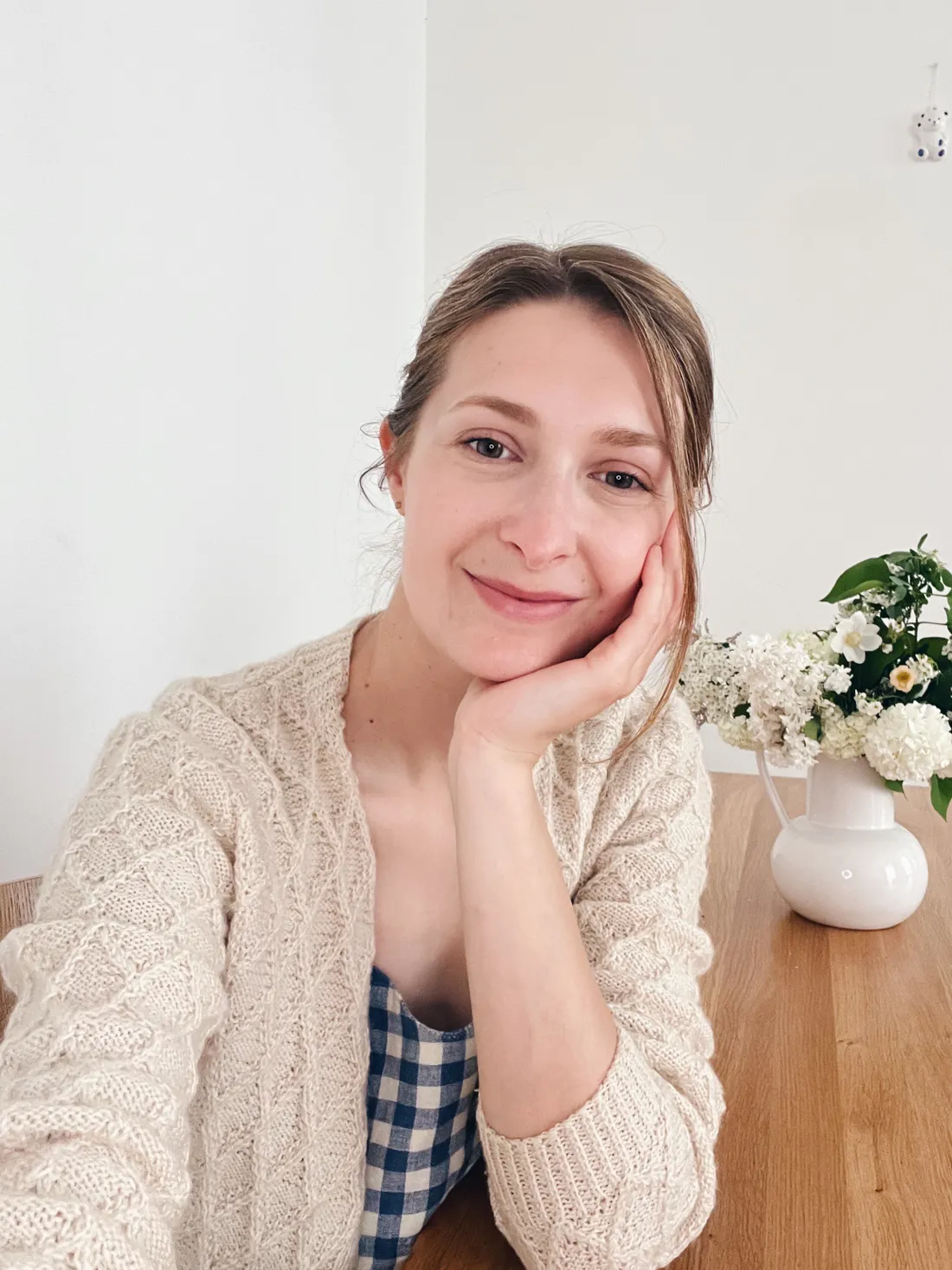 Žena v krémovém pleteném svetru a kostkované košili u dřevěného stolu, ruku podpírá bradu, vedle váza bílých květů.
