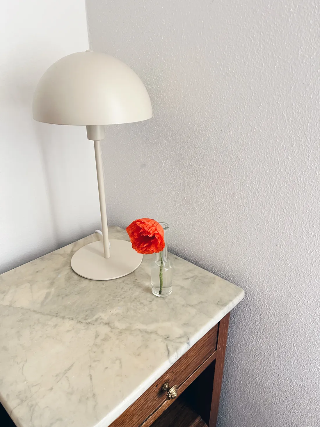 Krémová stolní lampa a oranžový mák ve skleněné váze na mramorovém nočním stolku u bílé stěny.