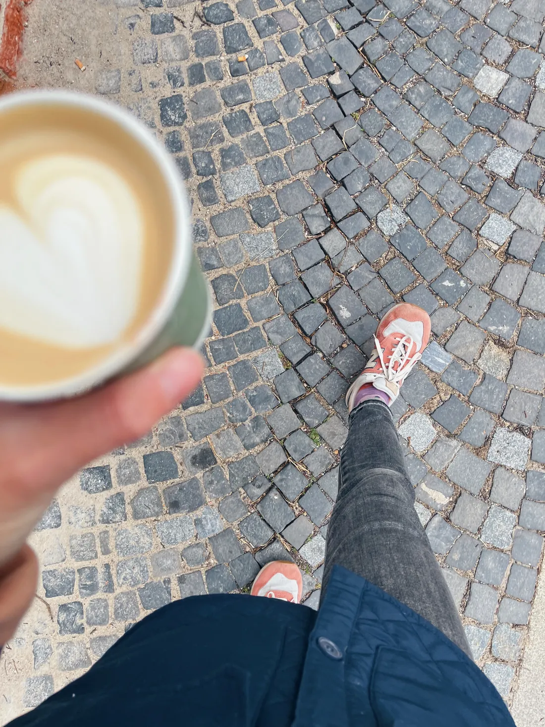 Pohled shora na ruku se šálkem cappuccina se srdcem, růžové tenisky a tmavé kalhoty na šedé dlažbě při chůzi.