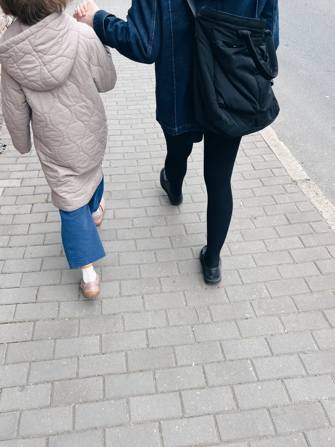Dospělý a dítě jdou za sebou po šedé dlažbě za ruce, dítě v béžovém prošívaném kabátu a modrých kalhotách.