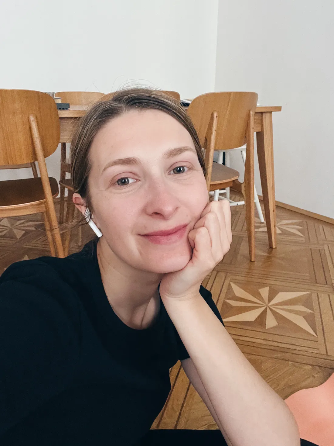 Žena v černém tričku se sluchátky se usmívá v jídelně s dřevěnými židlemi a parketami.