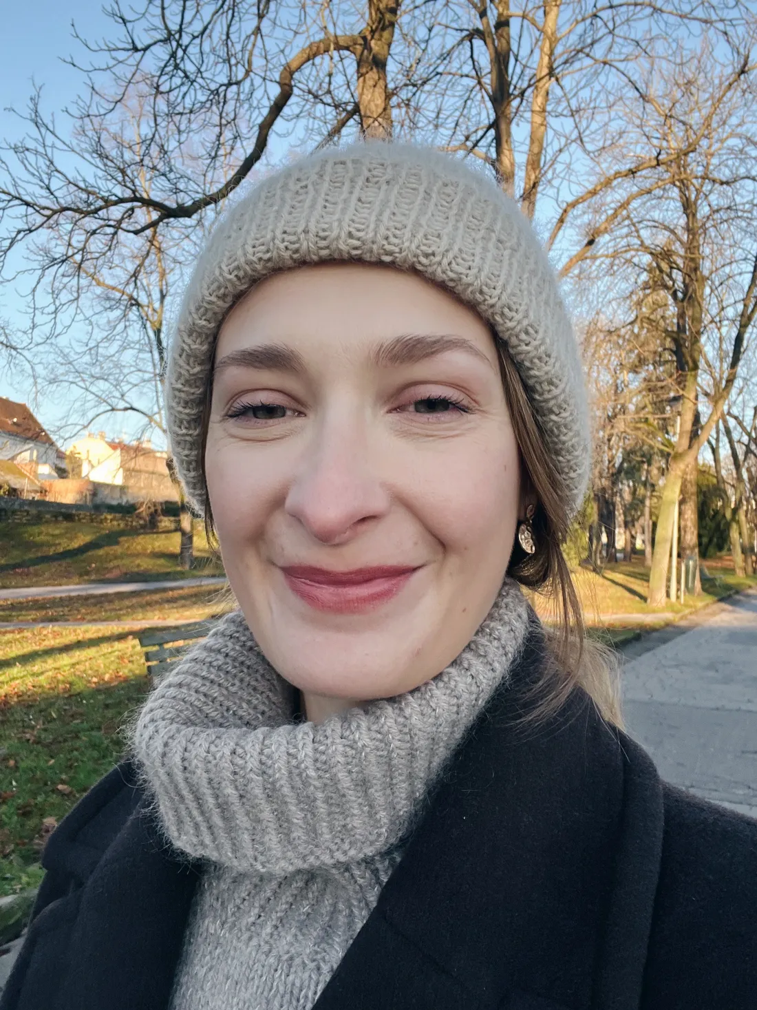 Usmívající se žena v béžové pletené čepici a roláku s černým kabátem v prosluněném parku se stromy.