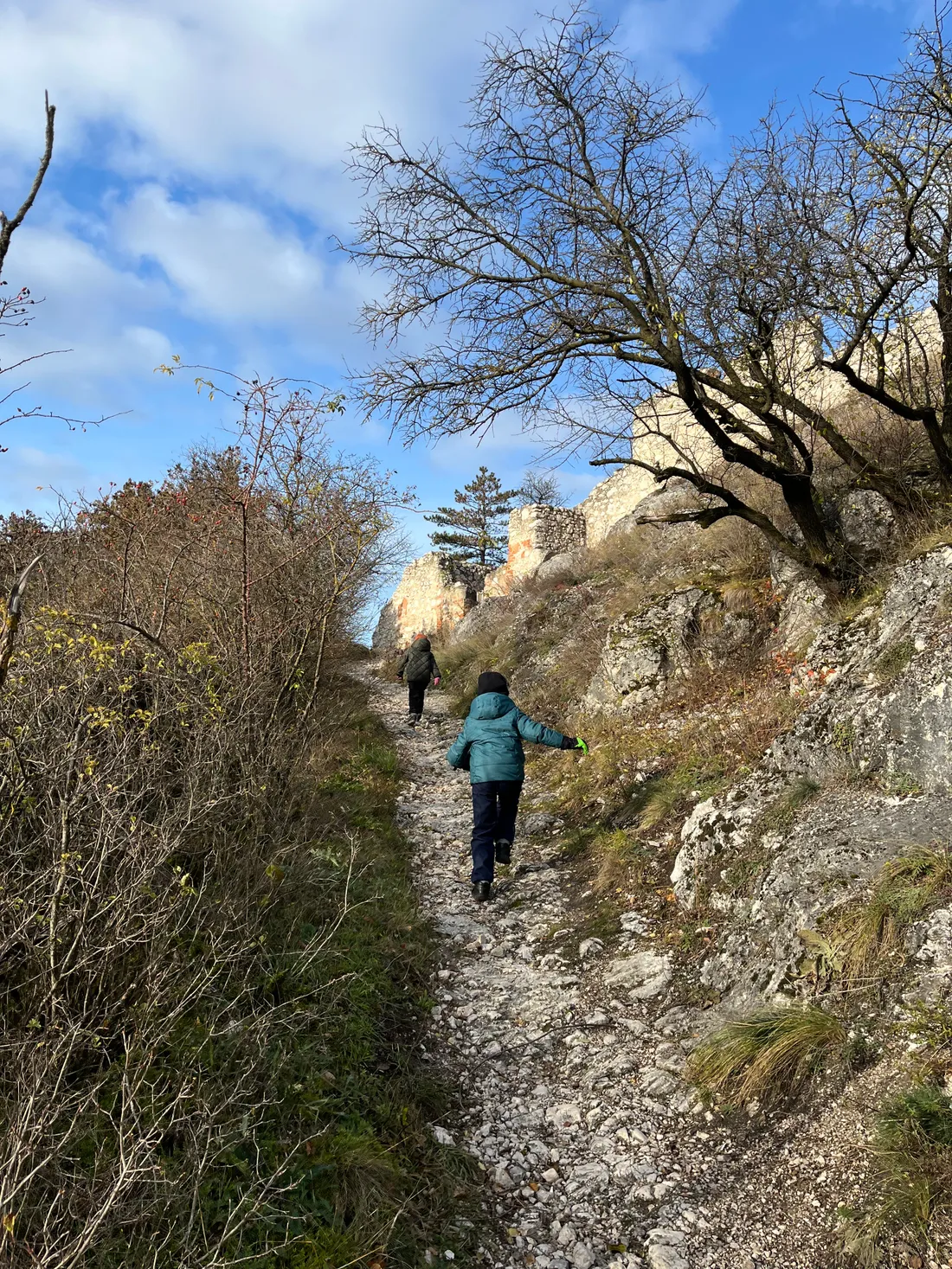 Dvě děti vycházejí po kamenné cestě na kopec k ruinám pod modrou oblohou.