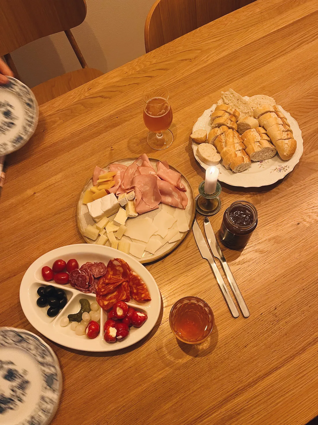 Dřevěný stůl se sýry, uzeninami, olivami, rajčaty, bagetou, pivem a zapálenou svíčkou.