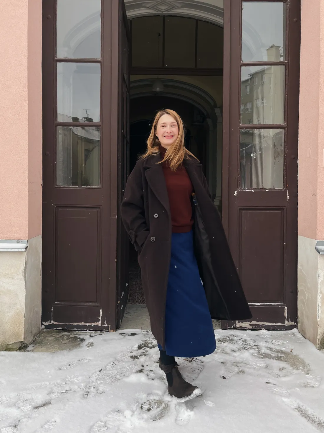 Usmívající se žena v hnědém kabátu a modré sukni před dřevěnými dveřmi ve sněhu.