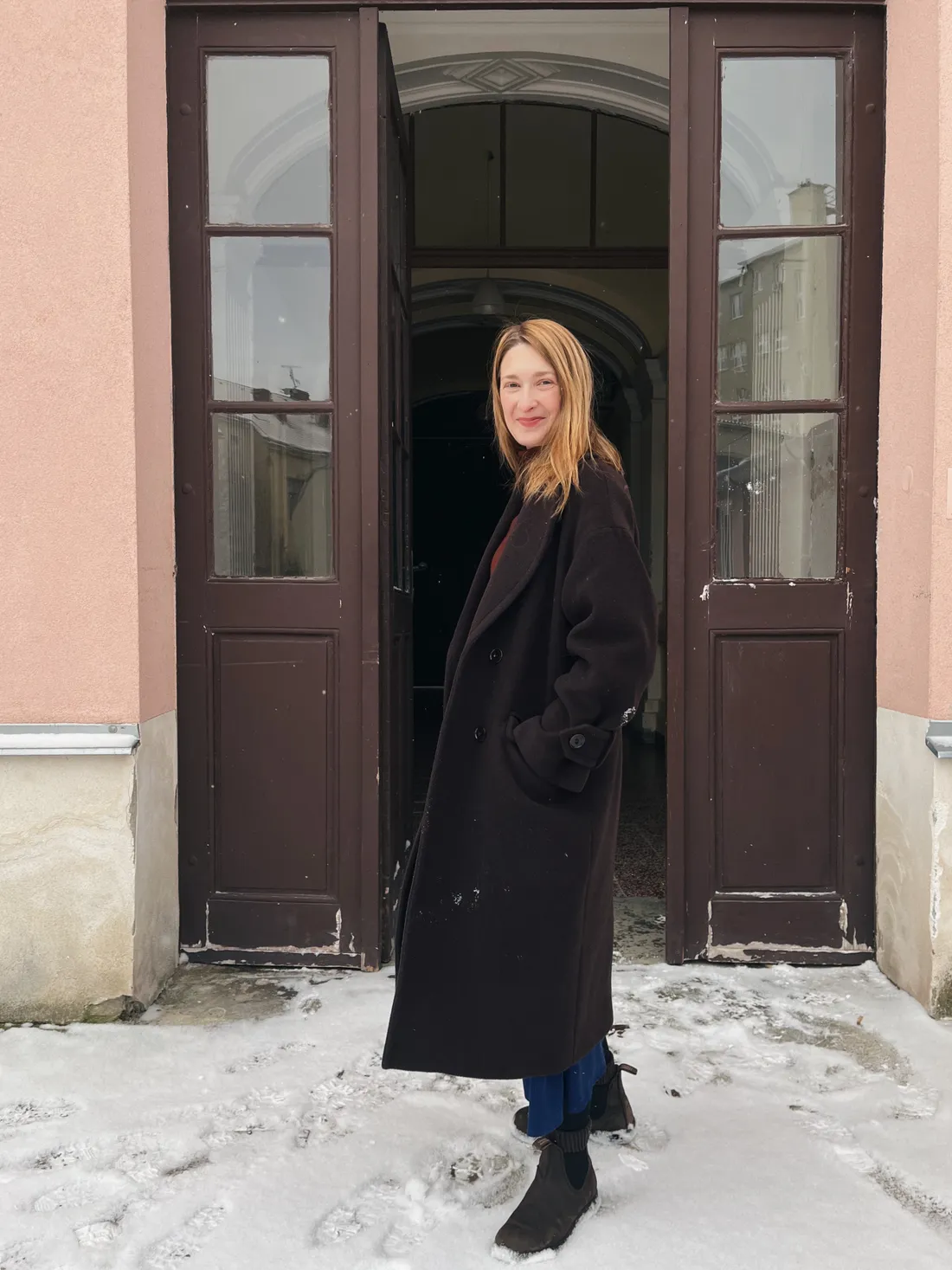Usmívající se žena v dlouhém tmavém kabátě stojí ve dveřích domu na zasněženém prahu.
