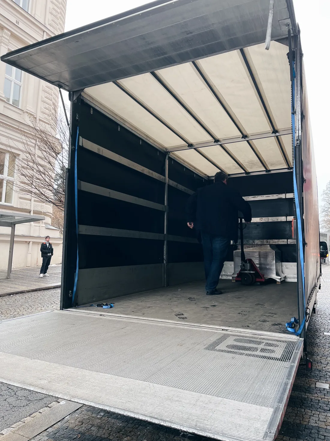 Muž v nákladním autě nakládá zabalený objekt na paletovém vozíku na rozkládací rampě.