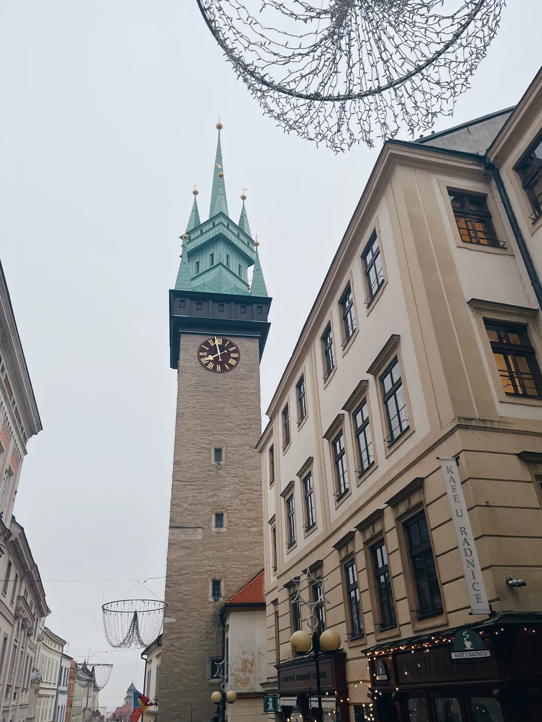 Vysoká kamenná radniční věž se zelenou střechou a závěsnými světelnými kruhy pod šedou oblohou.
