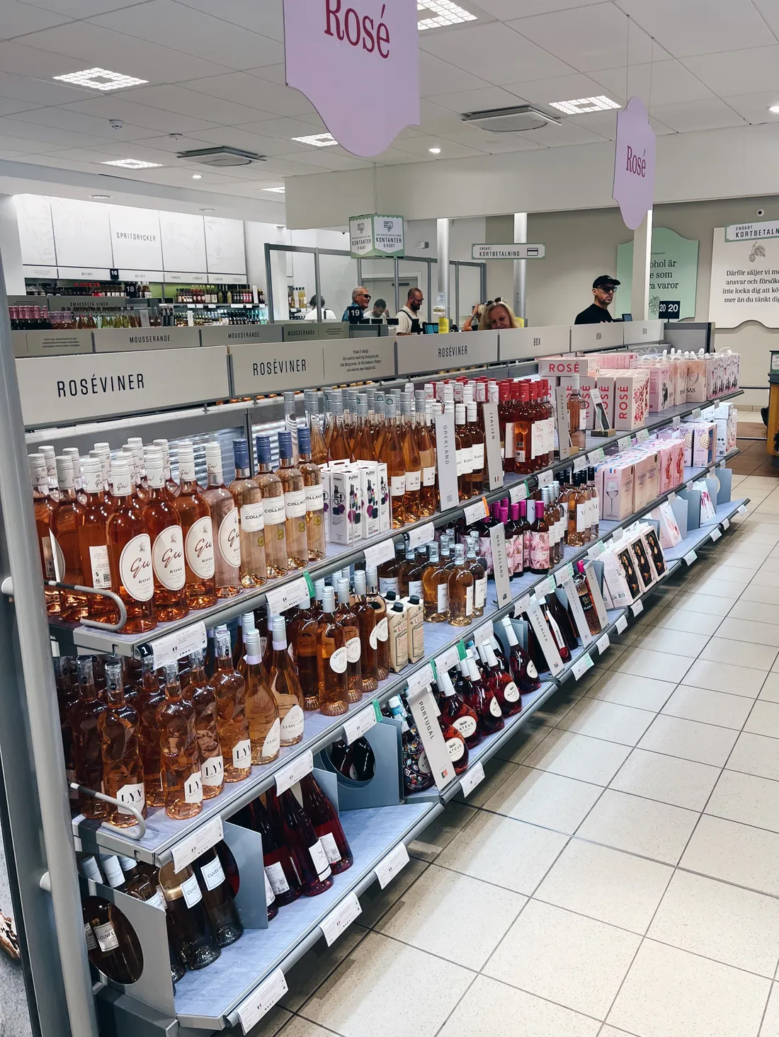 Regály plné lahví růžového vína s nápisy Roséviner a fialovými cedulemi Rosé ze stropu.