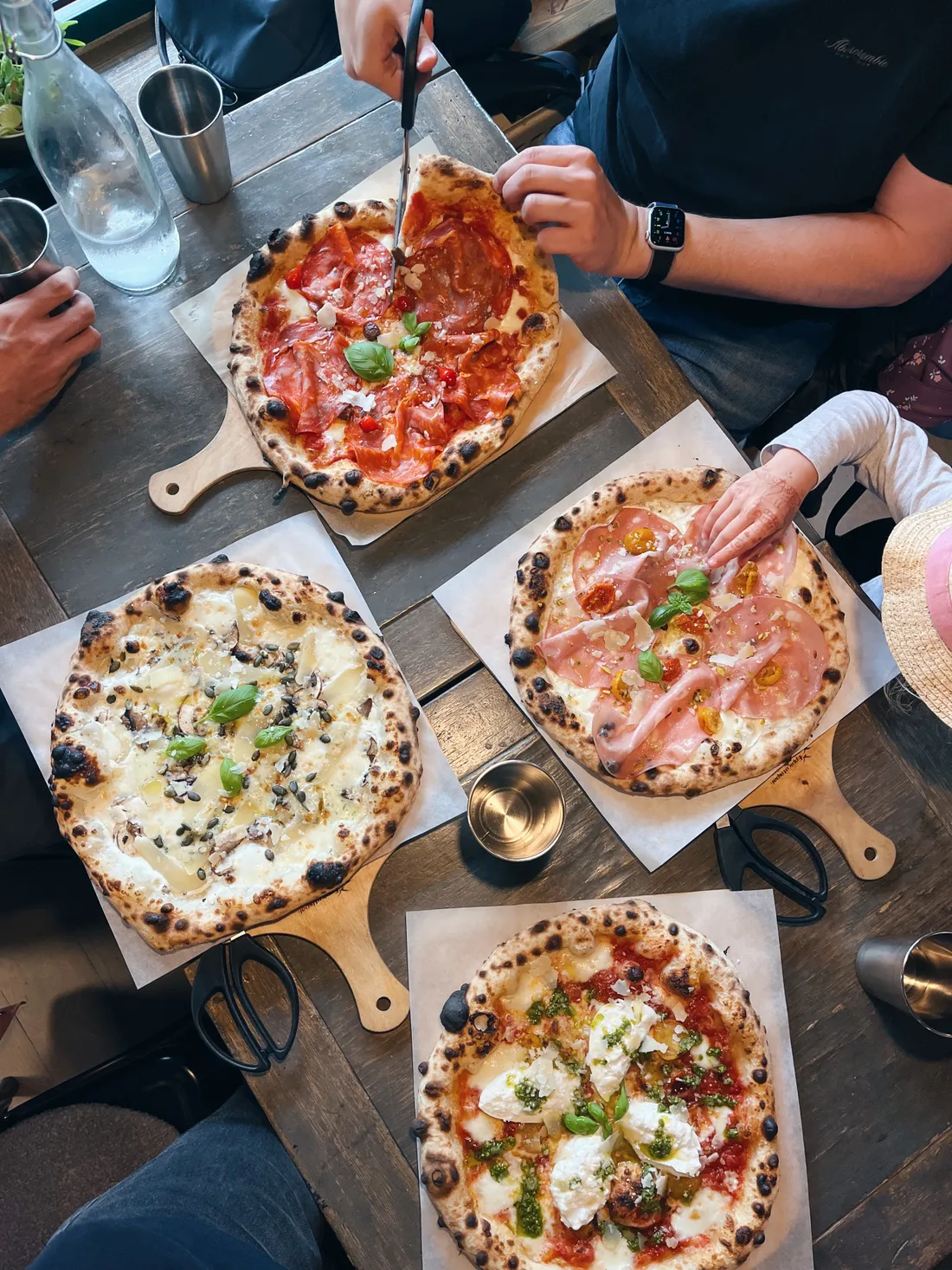 Čtyři neapolské pizzy na dřevěném stole, jedna se stříhá nůžkami, ruce stolujících.