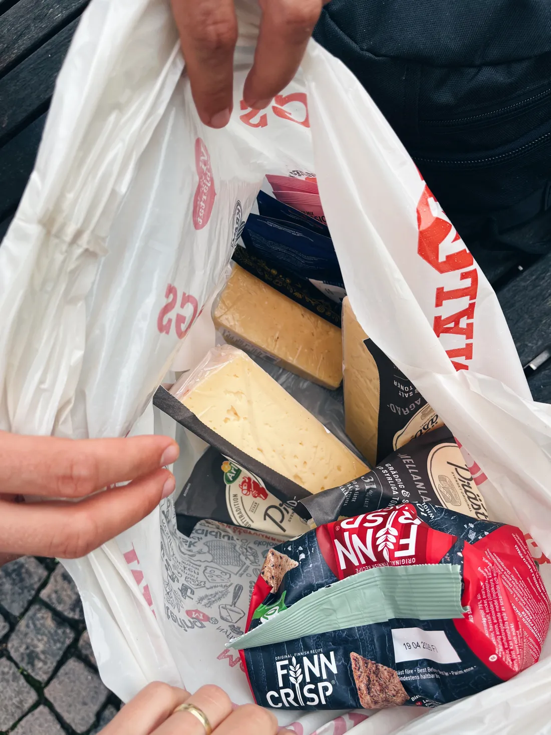 Ruce drží bílou nákupní tašku ICA se sýry, balením Finn Crisp a dalšími potravinami nad dřevěnou lavicí.