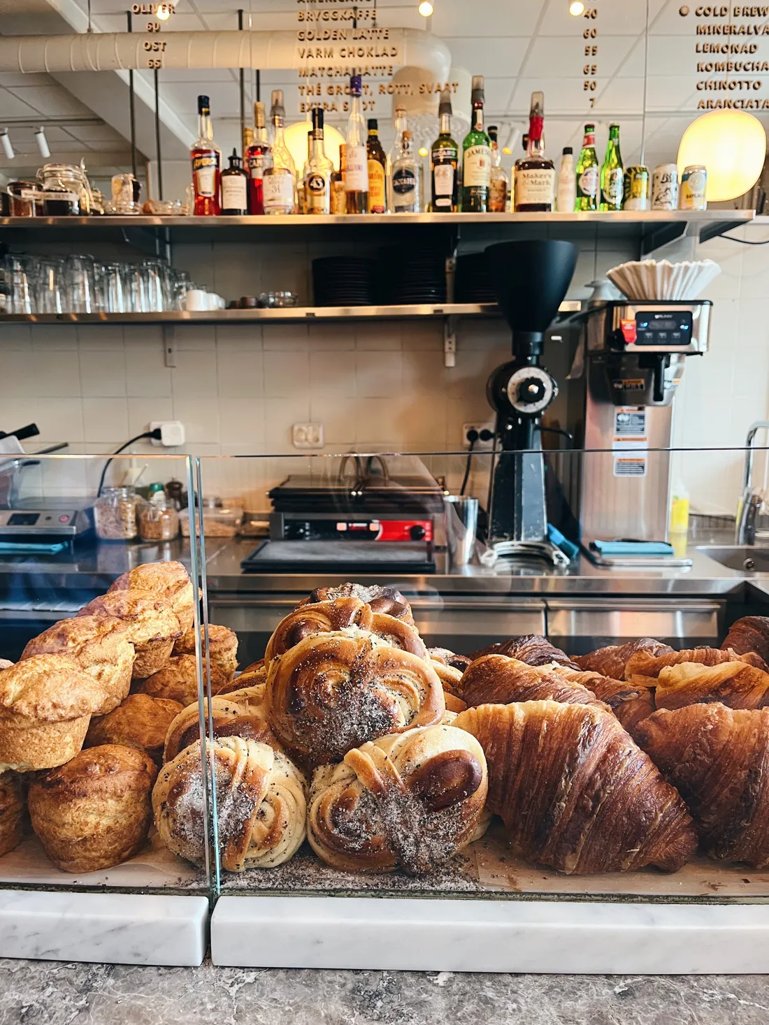 Vitrína s croissanty, šneky a muffiny u kávovaru a lahví v kavárně.