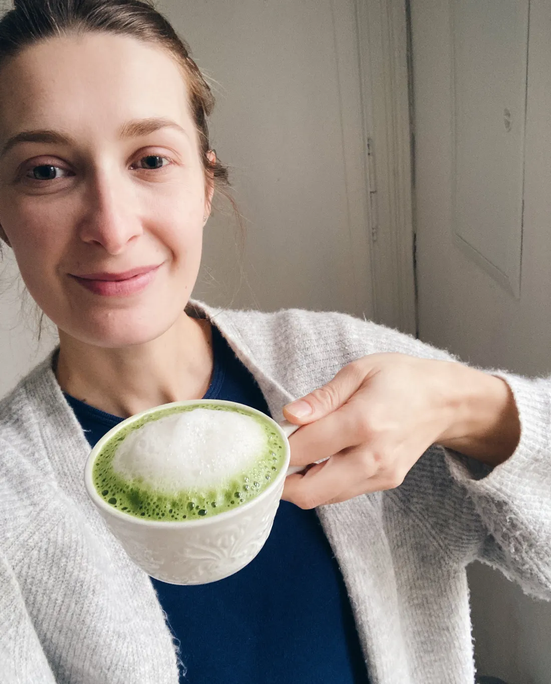 Selfie usmívající se ženy se šálkem zeleného matcha latte s bílou pěnou.