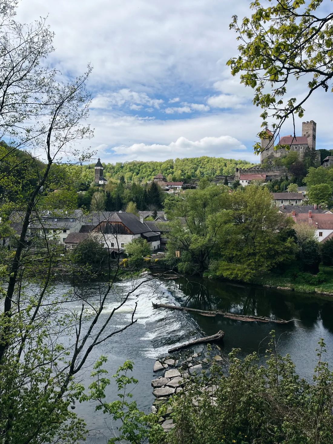 Řeka s přepadem, bílé domy a hrad na zalesněném kopci pod modrou oblohou s mraky.