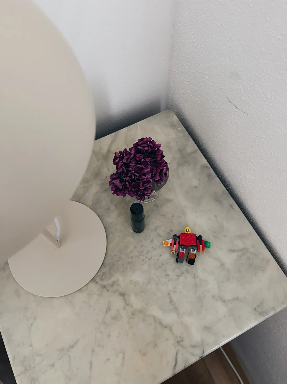 Mramorový stolek v rohu s vázičkou fialových květin, černou tyčinkou a červenočerným lego robotem.