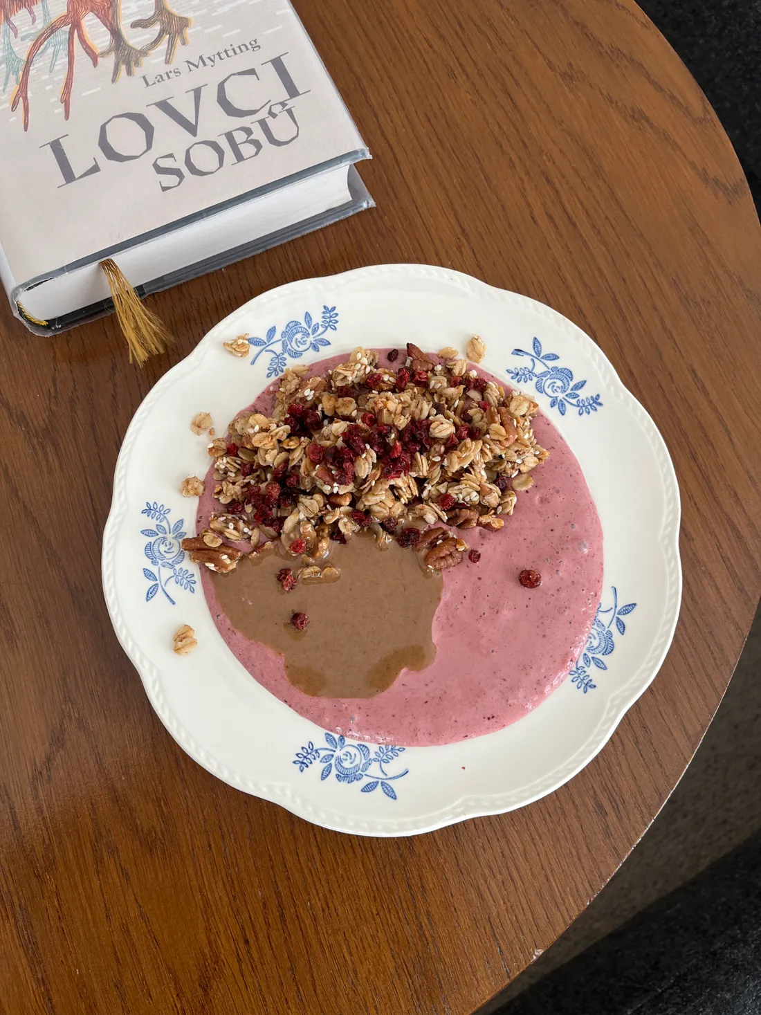 Růžová smoothie miska s granolou, ořechy a máslem na talíři s květinovým vzorem vedle knihy Lovci sobů.