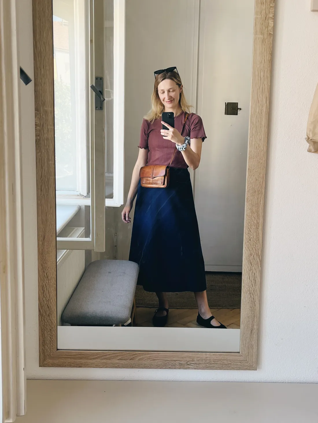 Selfie ve zrcadle: žena v malinovém tričku a tmavě modré sukně s koženou crossbody taškou a balerínkami.