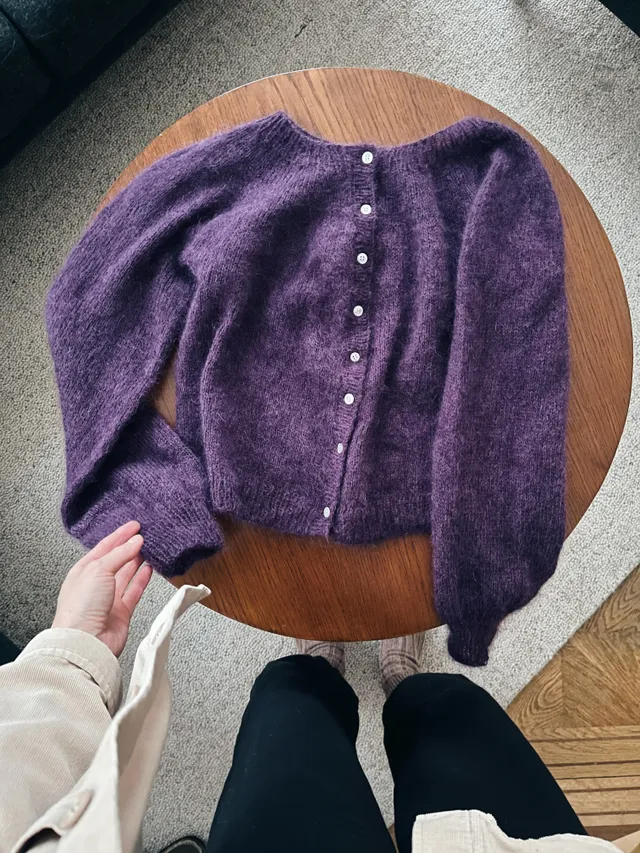 Huňatý fialový pletený kardigan s bílými knoflíky na kulatém dřevěném stole, ruka u manžety.