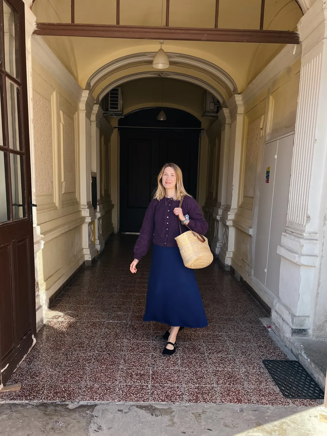 Žena v fialovém kardiganu a modré sukni se slaměnou taškou v klenuté chodbě.