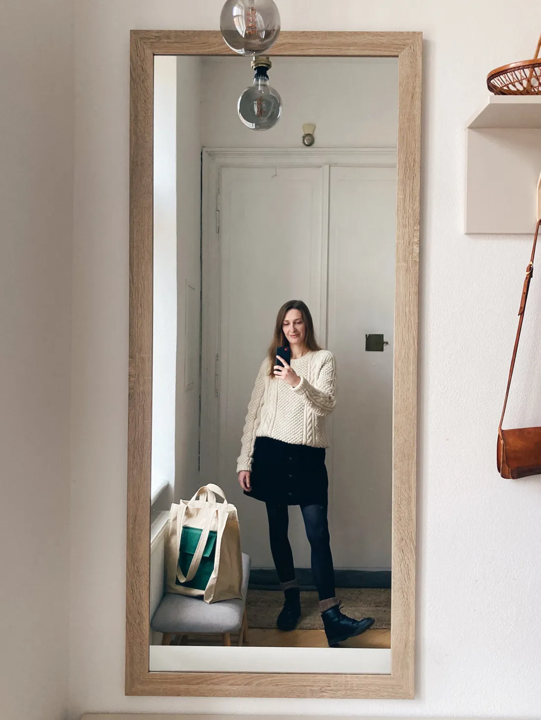 Selfie ve zrcadle: žena v krémovém pleteném svetru, černé sukni, punčochách a černých botách, proutěná taška u okna.