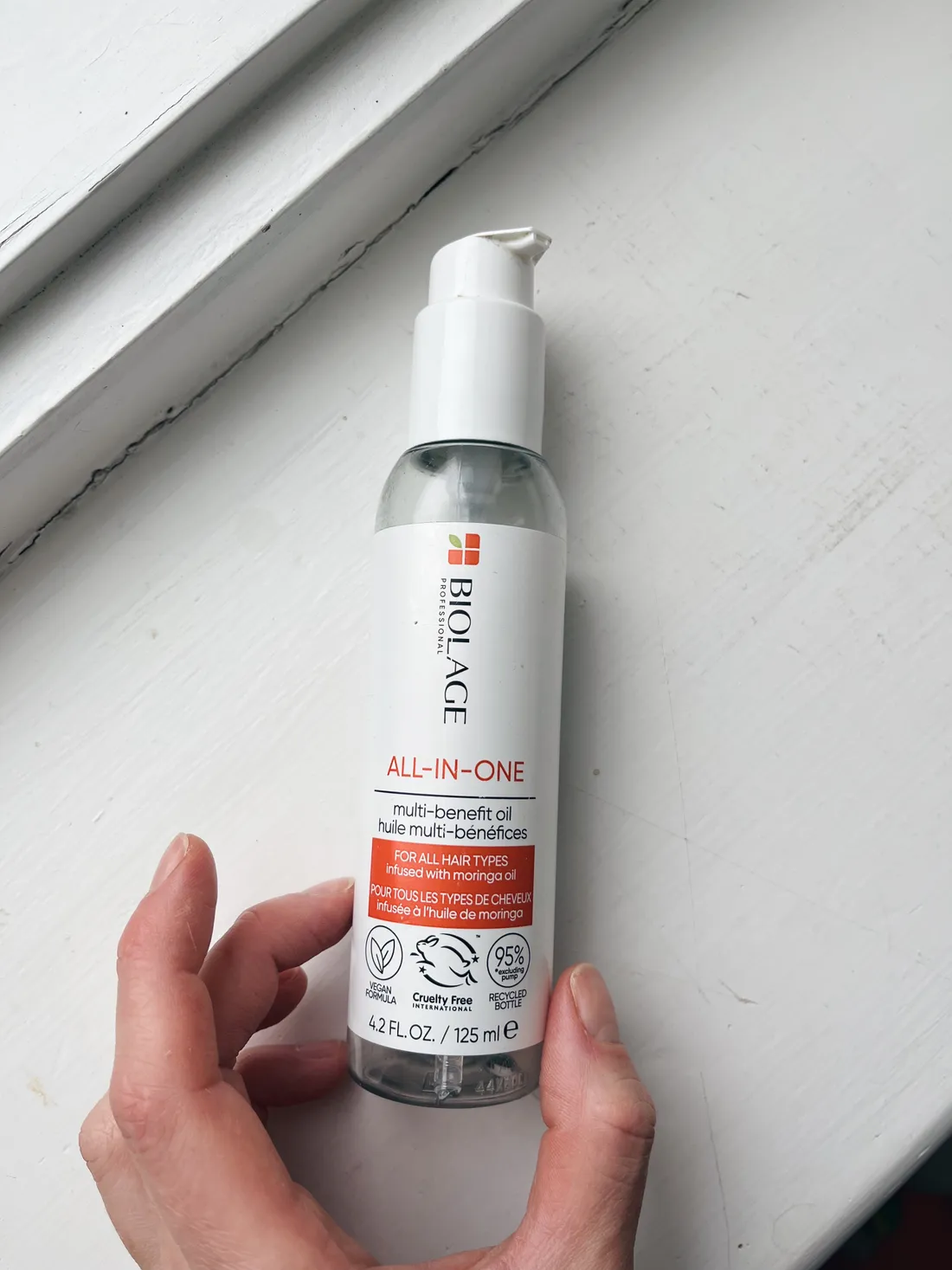 Lahvička oleje Biolage All-in-One s moringou v ruce na bílém dřevěném parapetu.